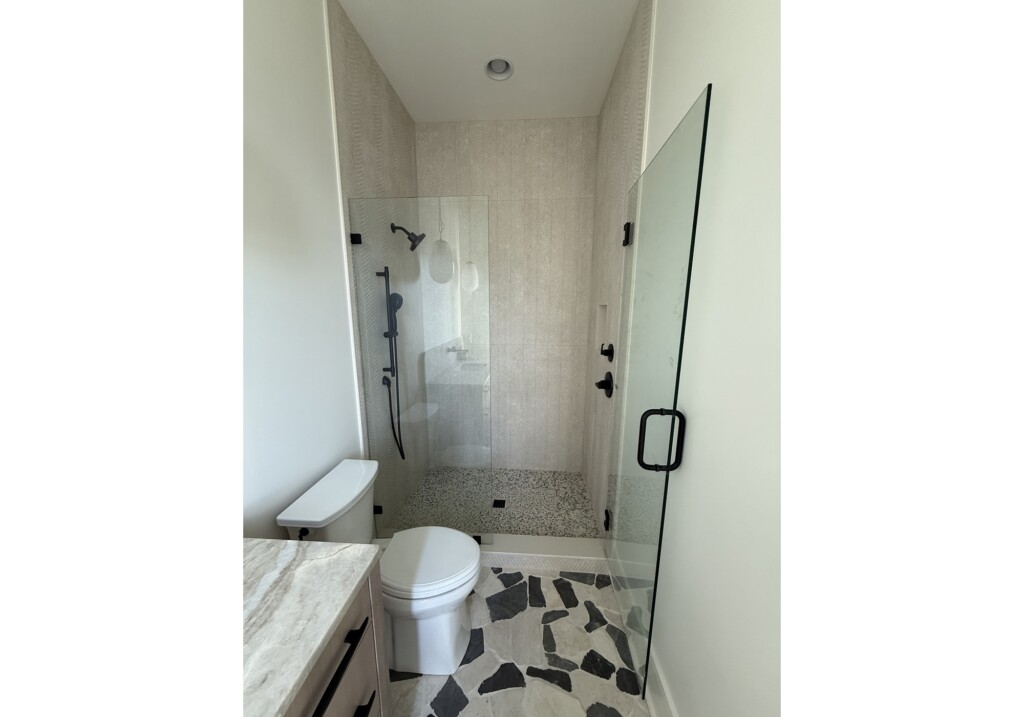 Woodward Project glass walk-in shower with white tile, black fixtures and pebble floor